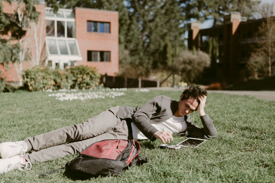 One student laying in the grass outside of an academic building, looking intently at an electronic device.