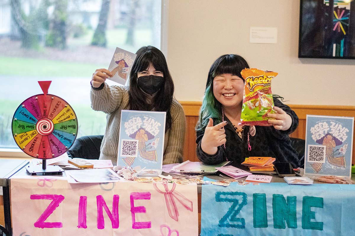 Two smiling librarians staff a promotional table filled with zines, games, and snacks