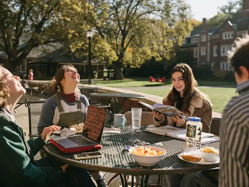 Four Reed College students sit around a round table outside. Two Reedies are laughing and one student looks into the camera with a knowing smile