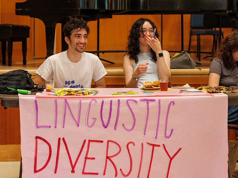 Two Reed College students sit at a table with a sign that says Reed Linguistic Diversity Project. They are smiling and looking at something to the right. 