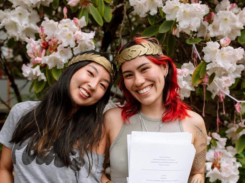 Two students wearing their new laurels and holding copies of their theses smile at the camera