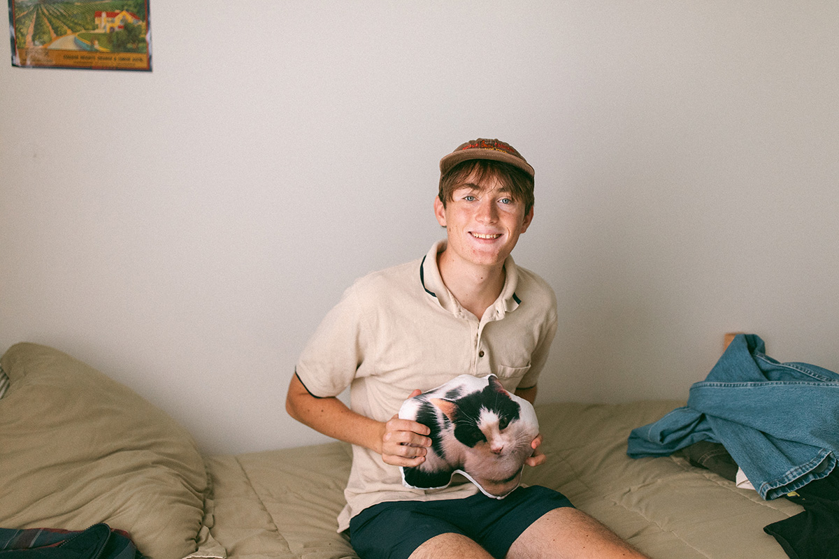 A student posing on thier bed with a small custom pillow of the family cat