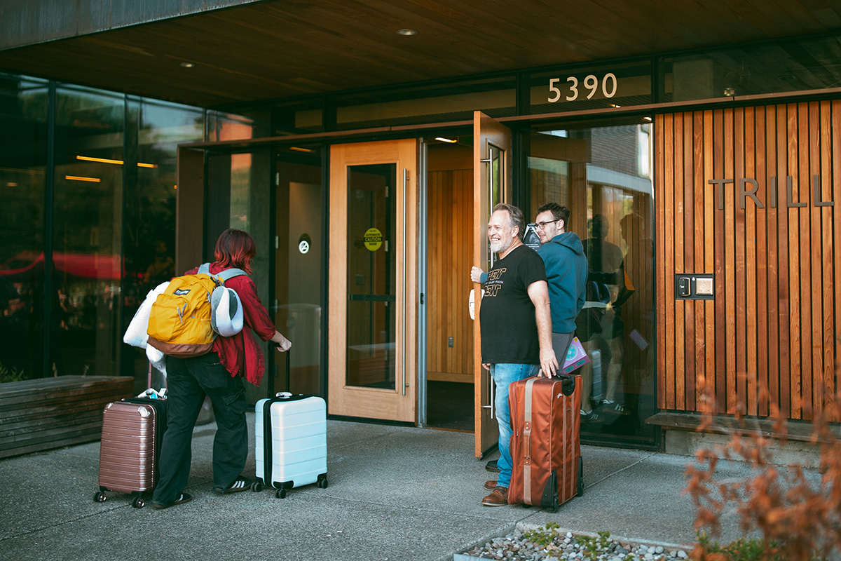 A family holding open the entrance door to Trilliam residence hall for a student pulling suitcases in