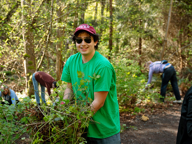 A student in a Reed baseball hat and green shirt holds up a pile of invasive ivy. Three students are working on the trail behind them.