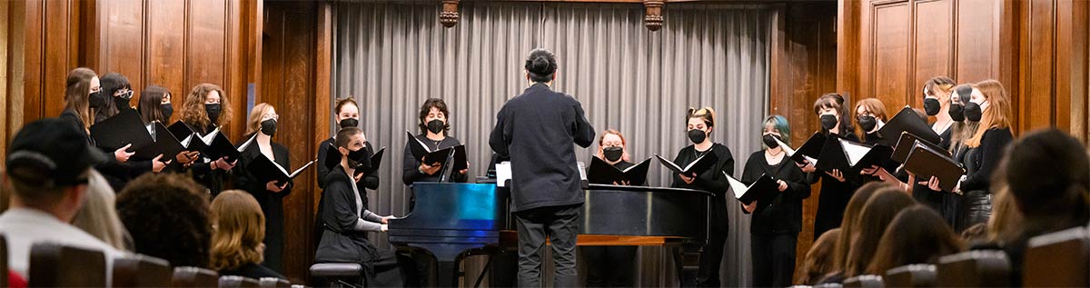 The Treble Voices Ensemble in performance, 16 singers surrounding a pianist