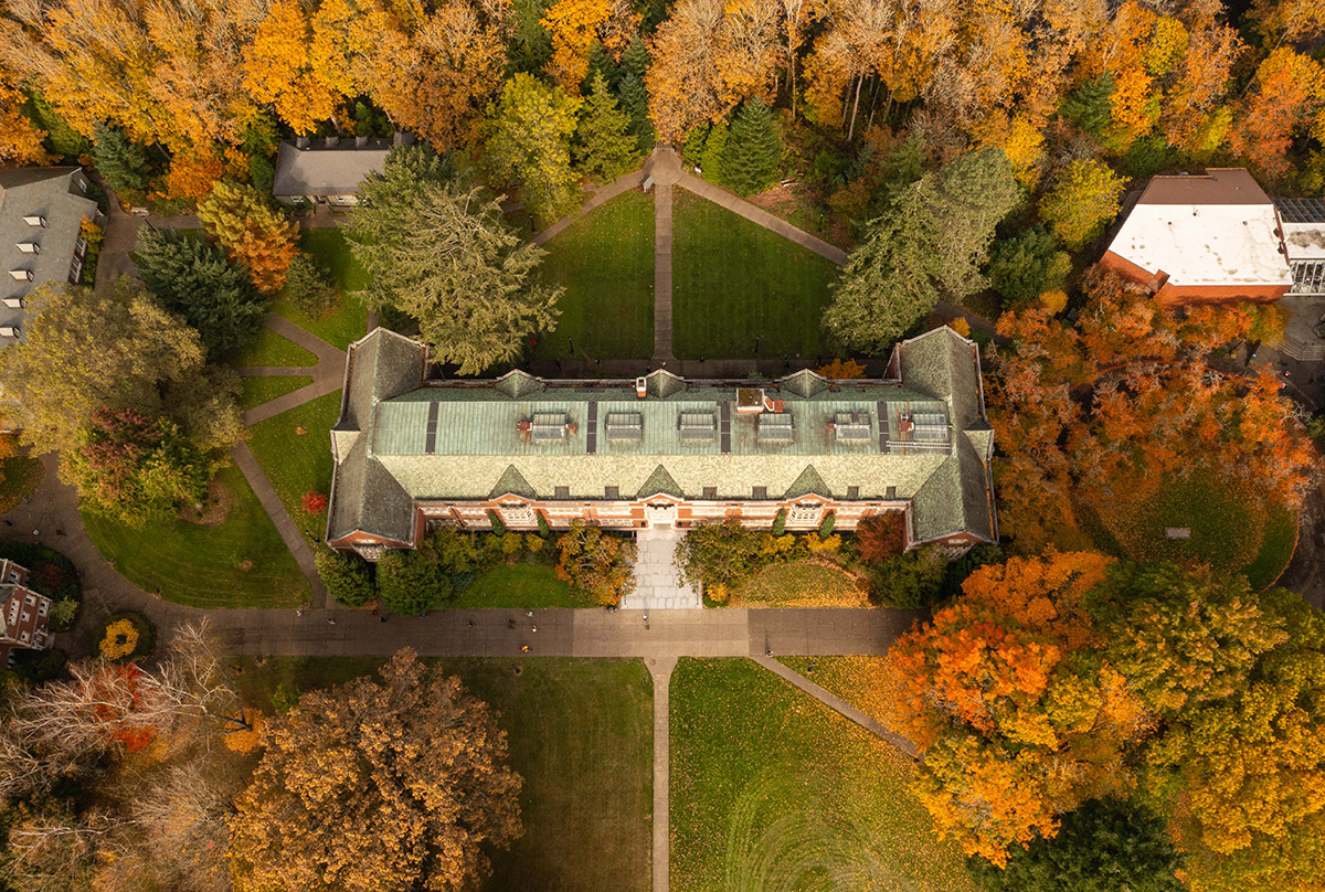 Aerial photo of Eliot Hall, a brick building surrounded by trees in autumn foliage, grassy lawns, and geometric walking paths.