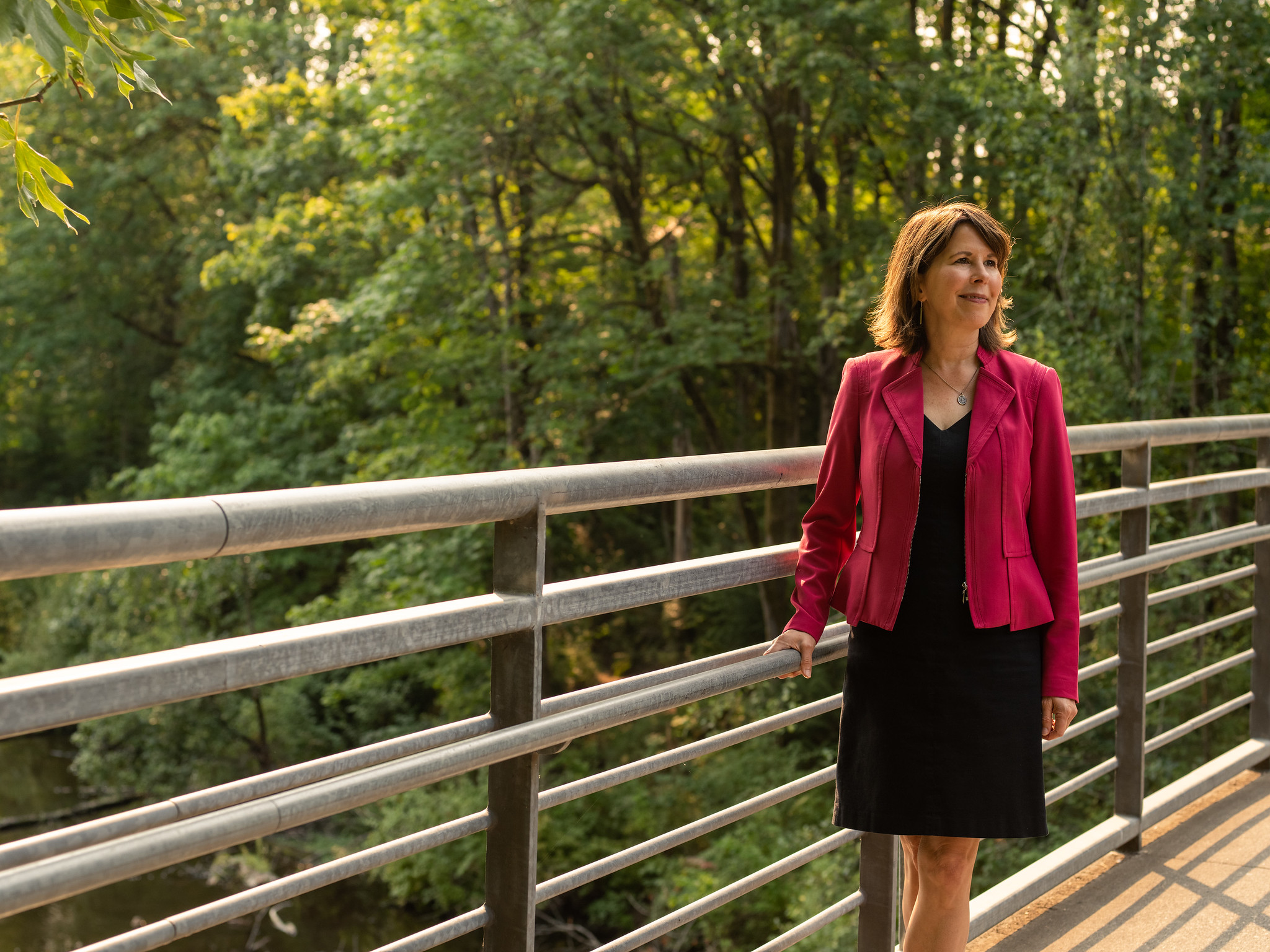 President Audrey Bilger, wearing a black dress and pink blazer, stands on a Reed College bridge, trees behind her.