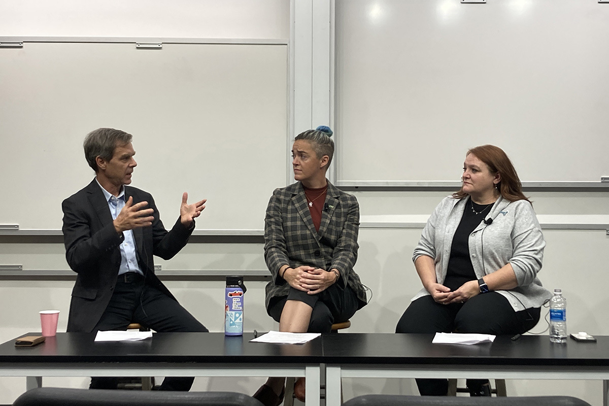 Three people sit behind a black table in front of whiteboards and hold a conversation.