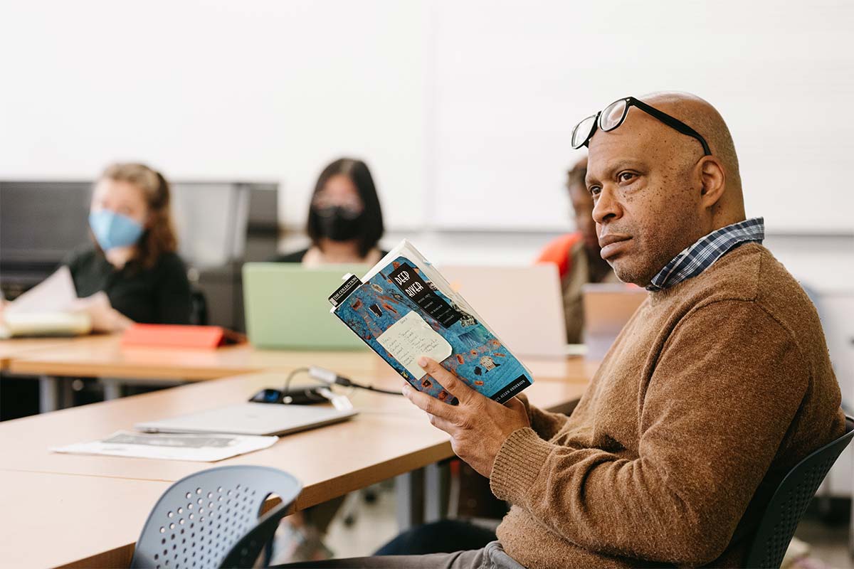 Professor of Music Mark Burford sits at a classroom table holding a book titled "Deep River: Music and Memory in Harlem Renaissance Thought"