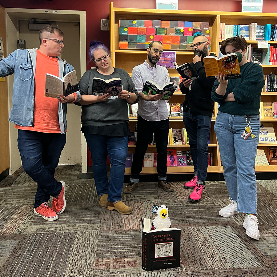 Five members of the bookstore staff reading books. They stand behind a plush griffin toy which is set in front of a copy of the book Comrades of the Quest, as if it is also reading.