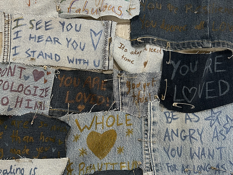 Rectangles of materials that have supportive messages written on them in fabric marker. All the pieces are held together with safety pins.